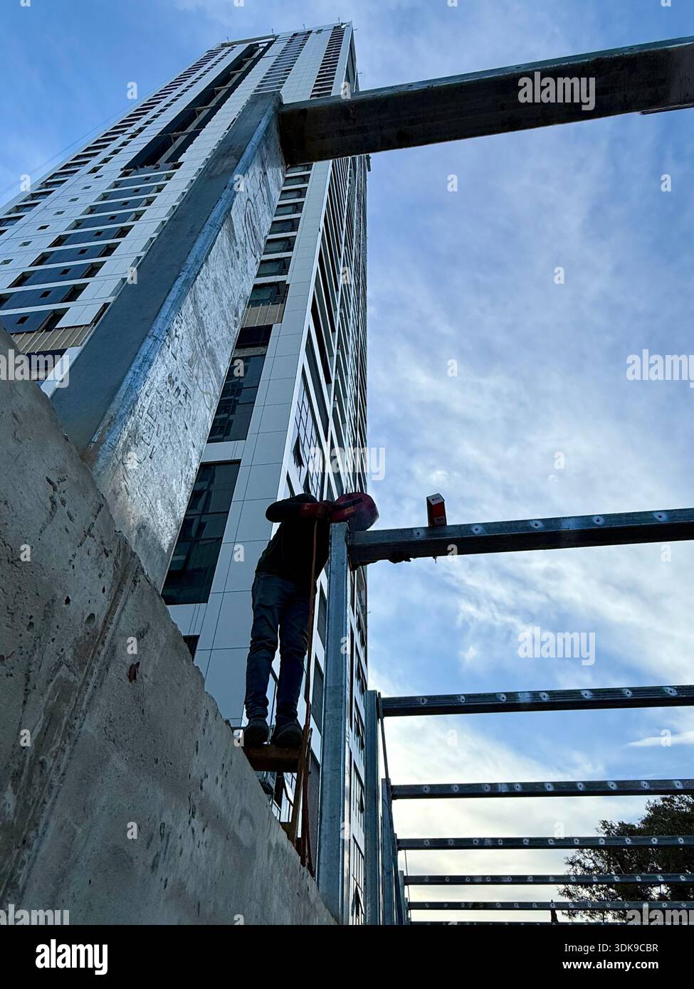 Construction worker on a ladder welding steel beams at a high-rise building site. Low-angle view of industrial labor against a modern skyscraper. - Smartphone Captured Stock Image