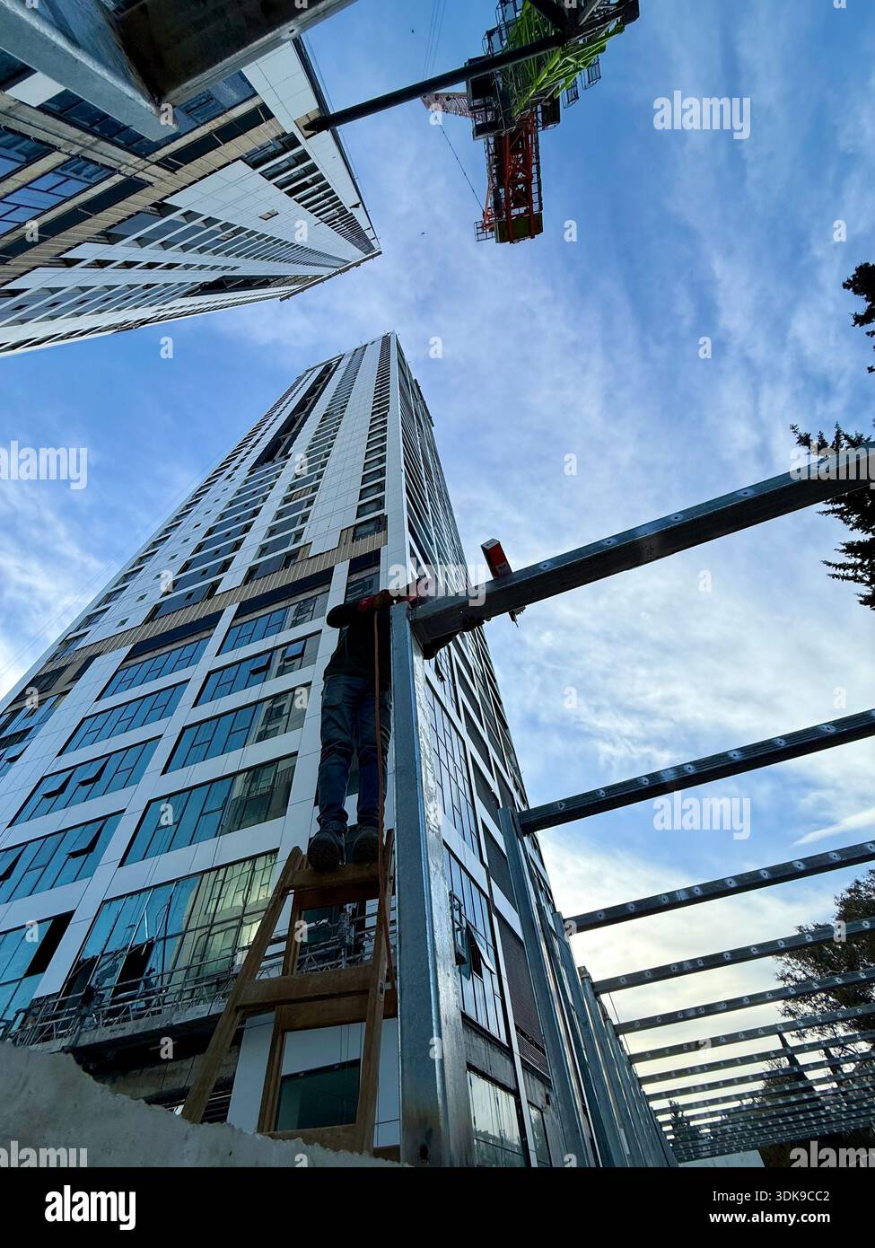 Construction worker on a ladder welding steel beams at a high-rise building site. Low-angle view of industrial labor against a modern skyscraper. - Smartphone Captured Stock Image