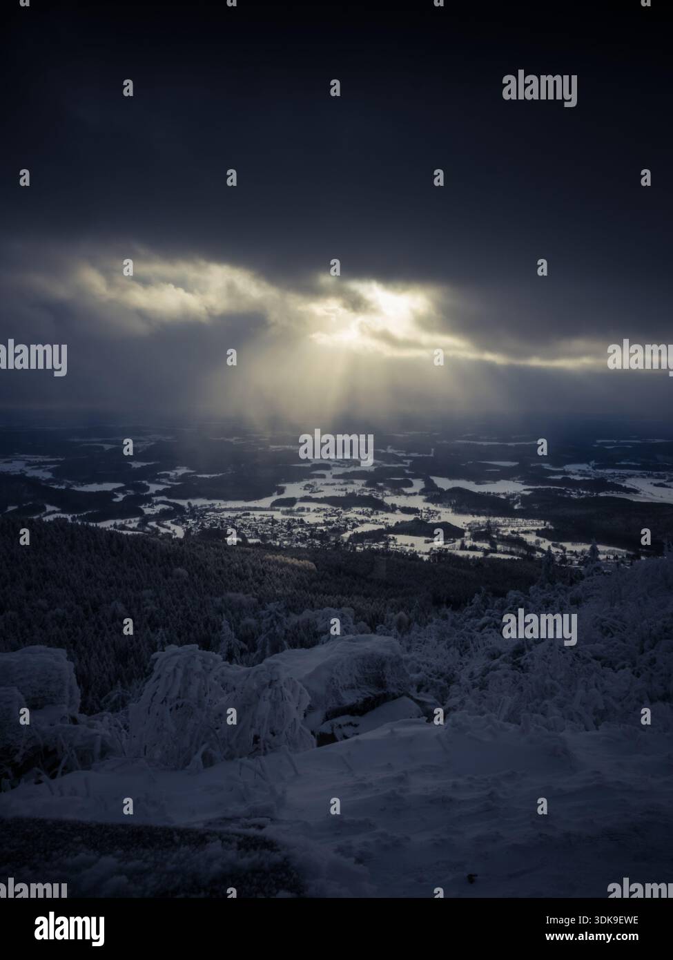 Sunbeams breaking through dark winter clouds above a snow-covered landscape viewed from Ještěd Mountain, creating a dramatic natural light display. Stock Photo