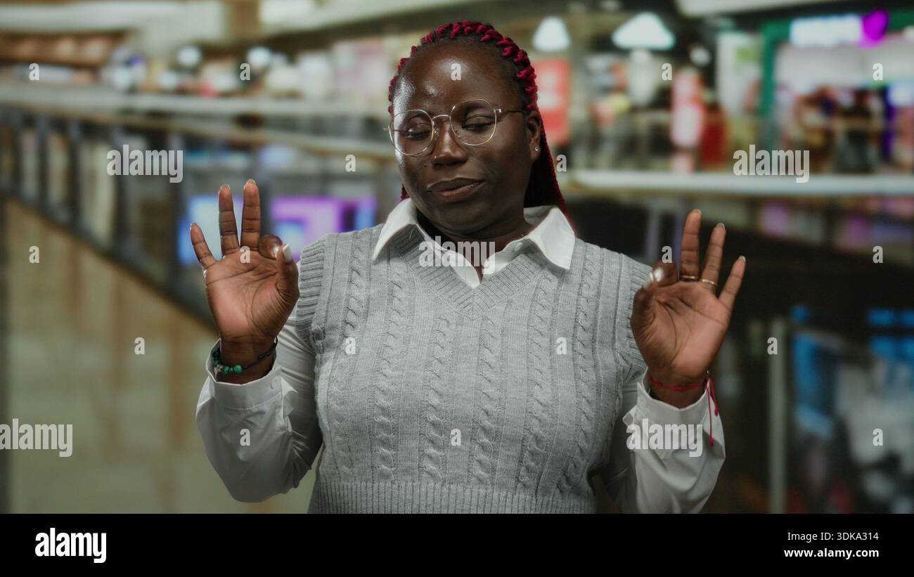 Woman meditating indoors in a crowded mall with people in the background showing a calm pose and serene expression with hands raised slightly in a ges Stock Photo