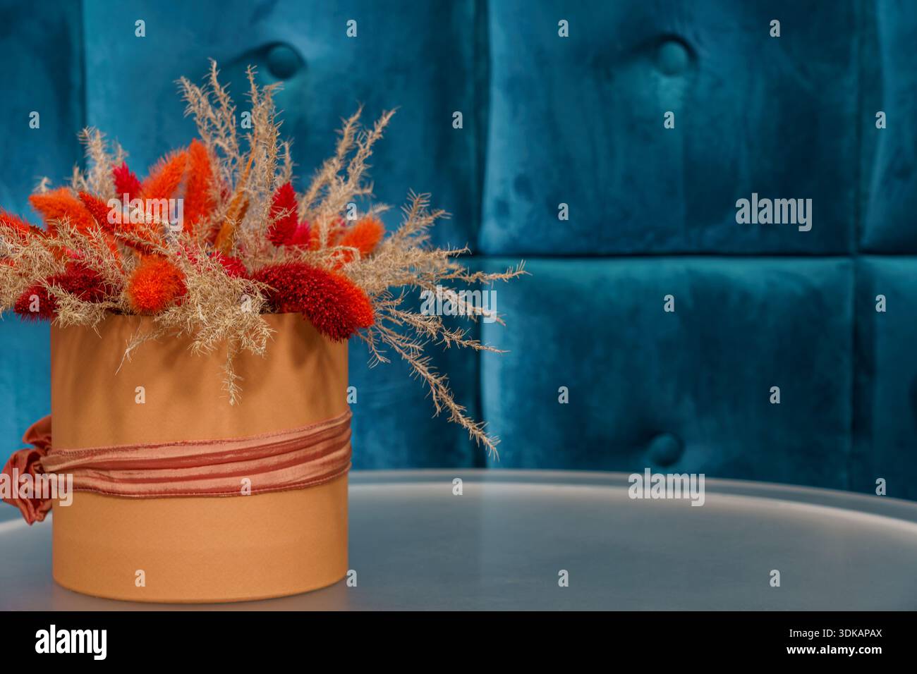 A flower arrangement with orange and beige tones sits in a pot on a table in front of a blue wall. Stock Photo