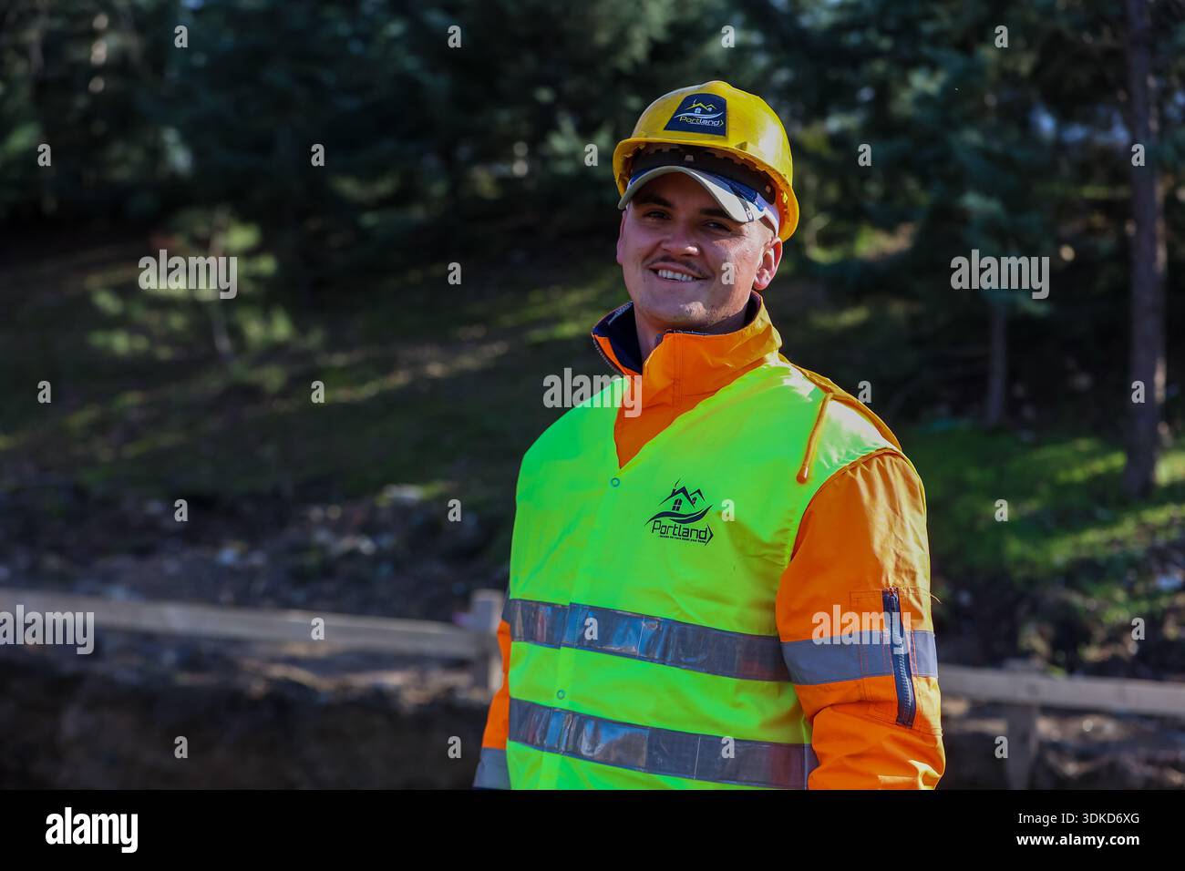 Smiling construction worker wearing a yellow safety helmet and high-visibility vest at an outdoor building site Stock Photo