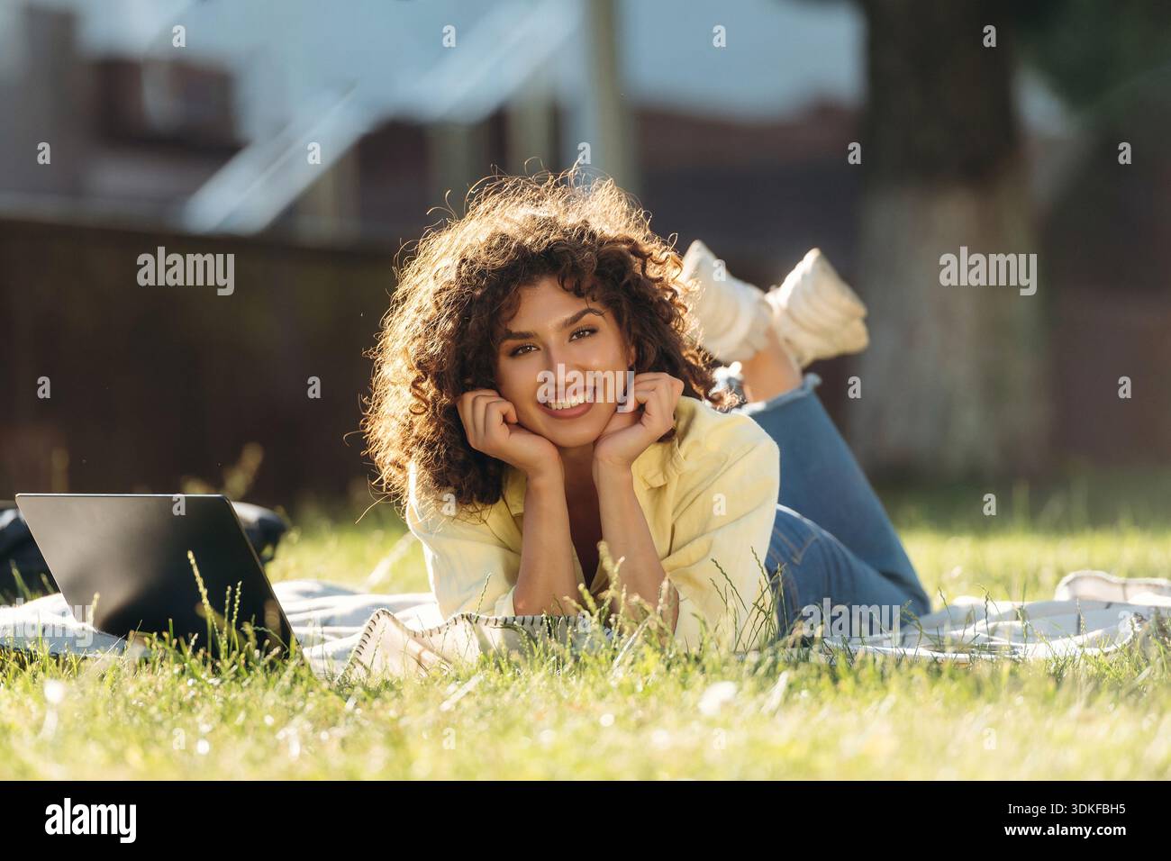 Smiling woman student is lying down on the green grass with laptop. Stock Photo