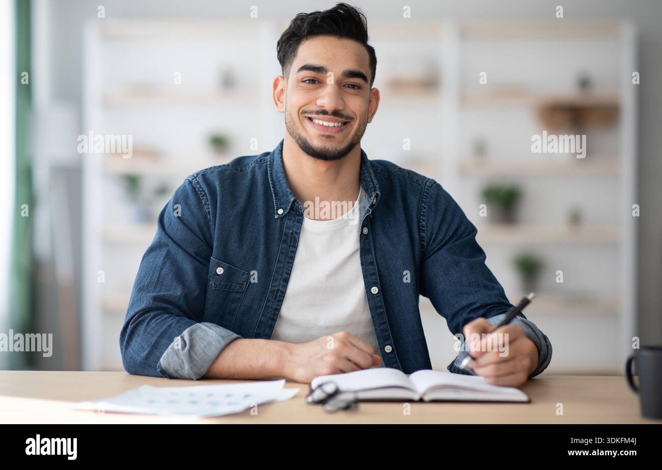Cheerful arab guy in casual sitting at workdesk, taking notes Stock Photo