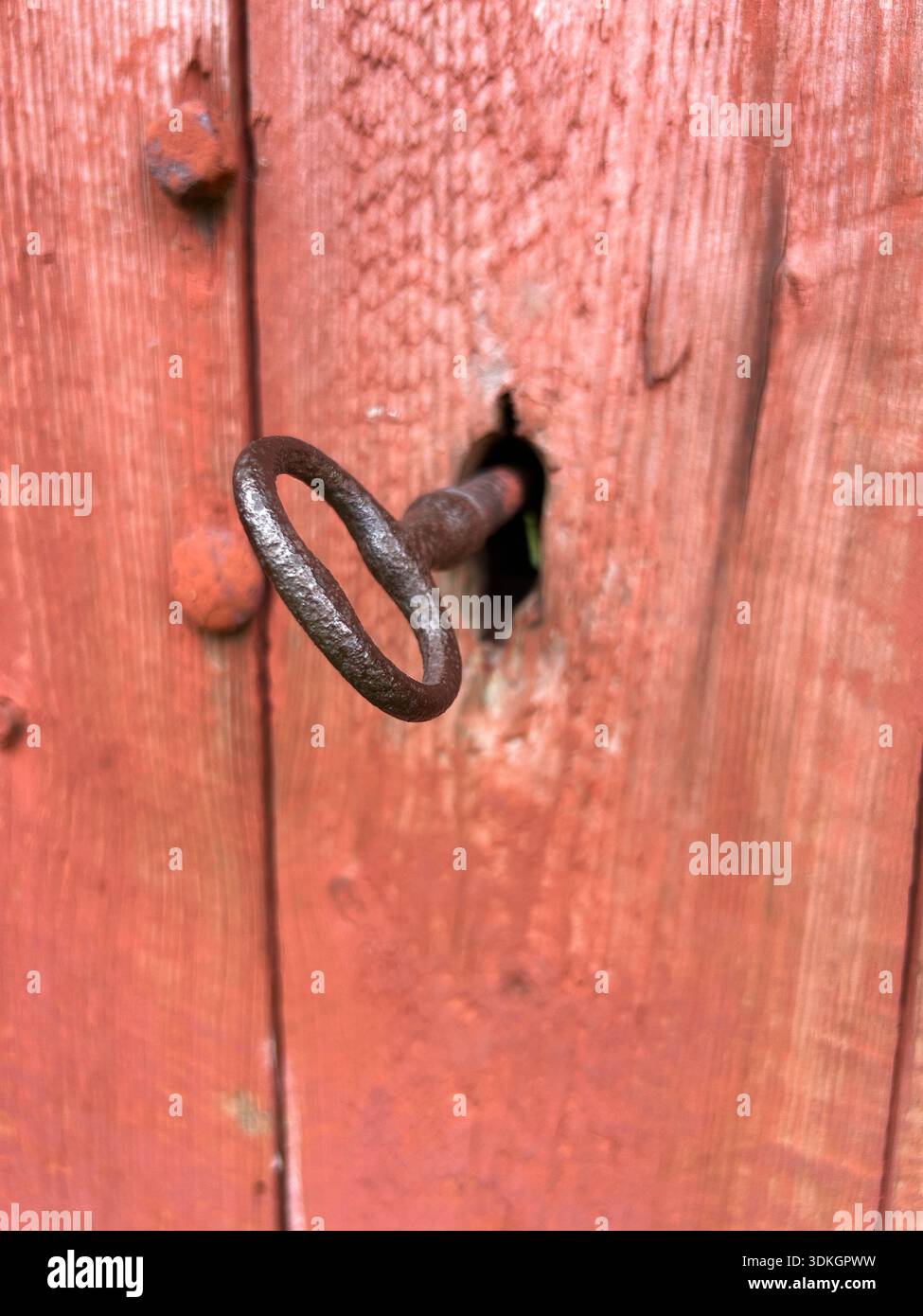 Close-up of a hand-forged key in a rustic keyhole on a red painted wooden barn door, Scandinavian rural detail. - Smartphone Captured Stock Image
