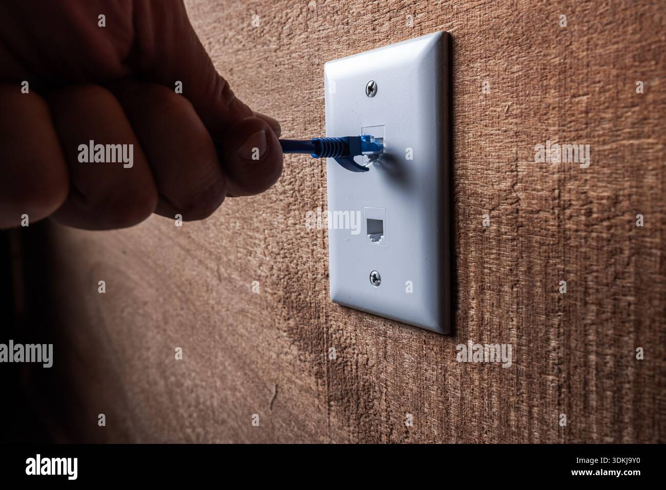 A close-up, high-detail shot of a hand plugging a blue RJ45 Ethernet cable into a white wall-mounted network port. Stock Photo