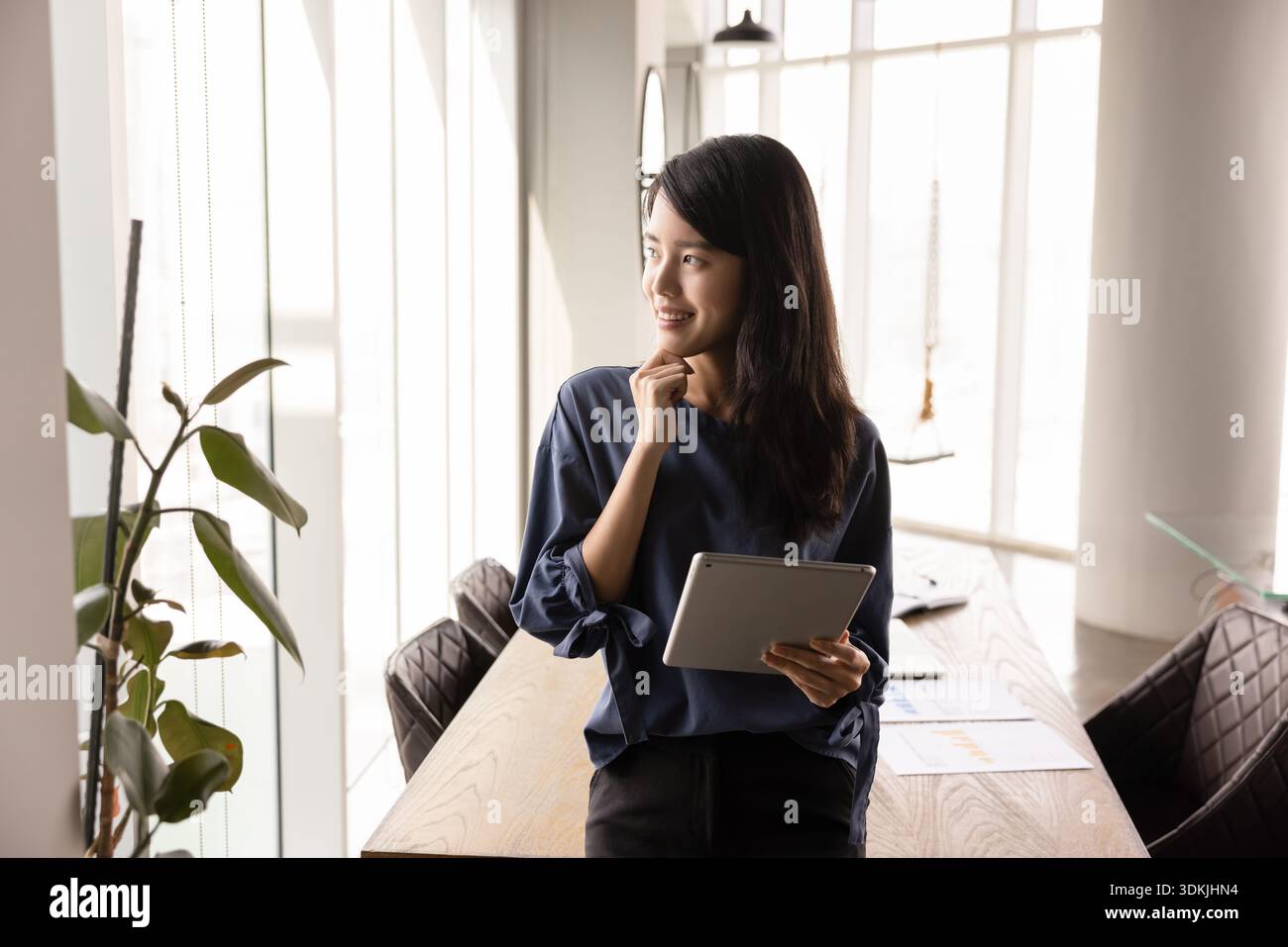 Happy thoughtful young Asian business leader woman holding tablet pc Stock Photo