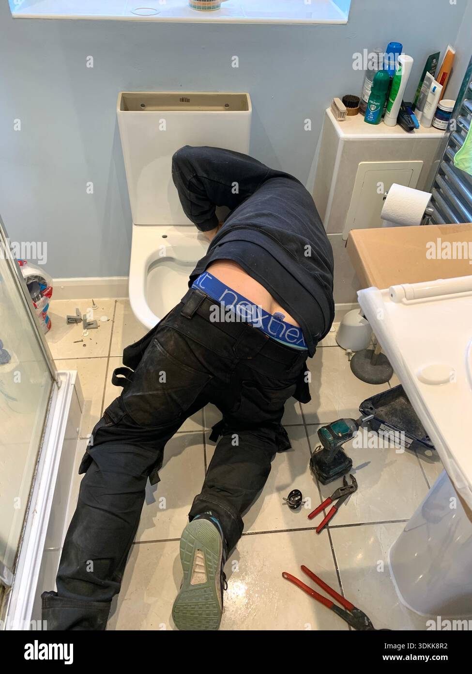 A person Kneeling on a bathroom floor while repairing a toilet with tools and fixings scattered on the tiles in a domestic bathroom floor Northampton - Smartphone Captured Stock Image