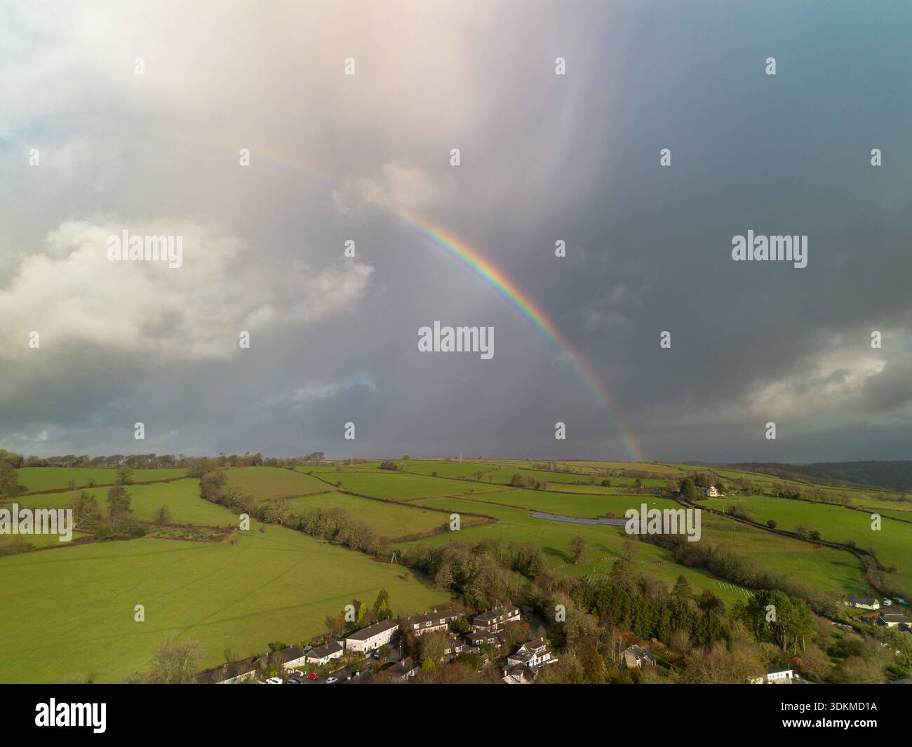 Bright rainbow over green countryside fields and a rural village under dramatic storm clouds. Stock Photo