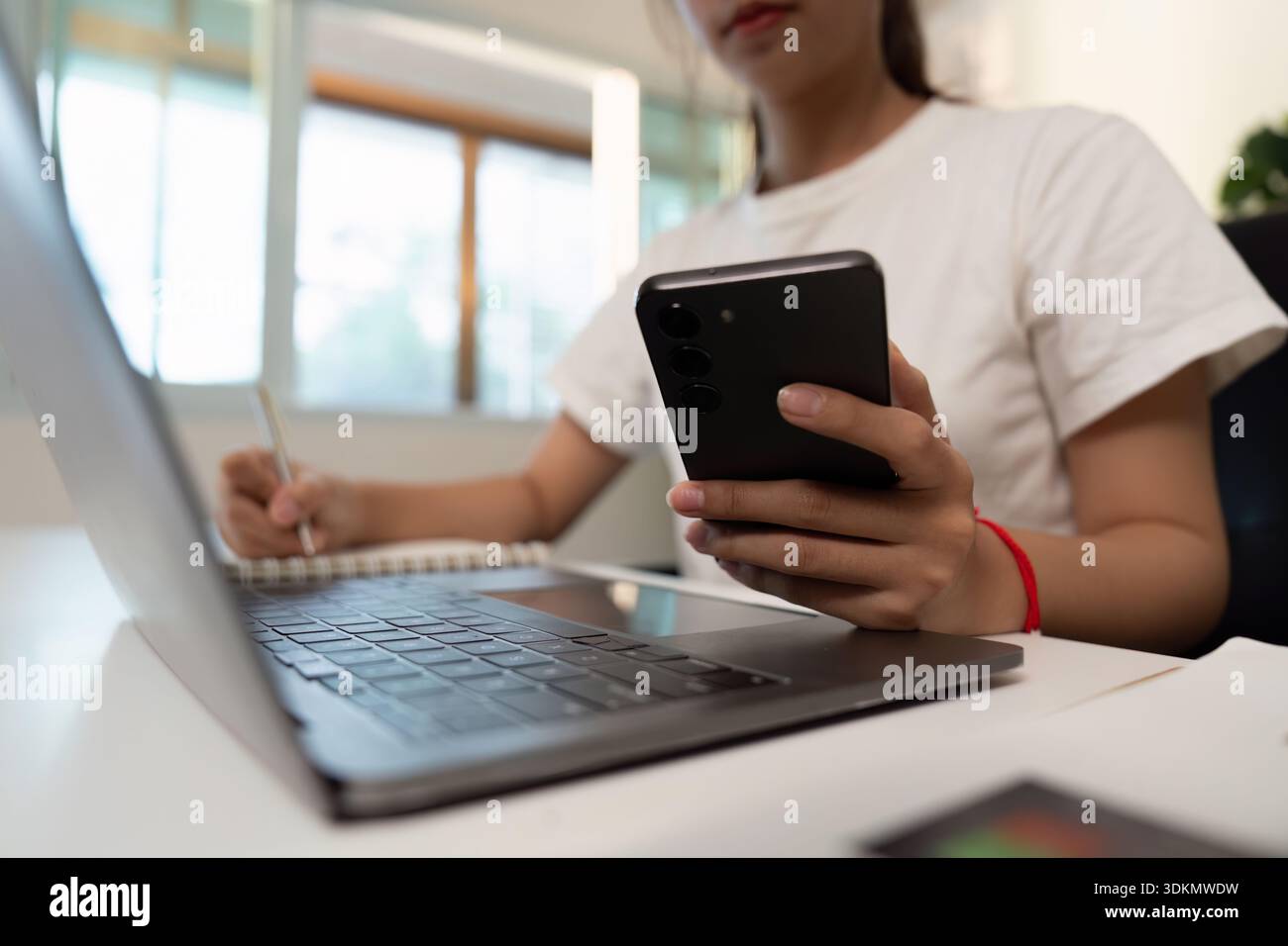 Multitasking. Young individual using smartphone while taking notes on laptop. Stock Photo