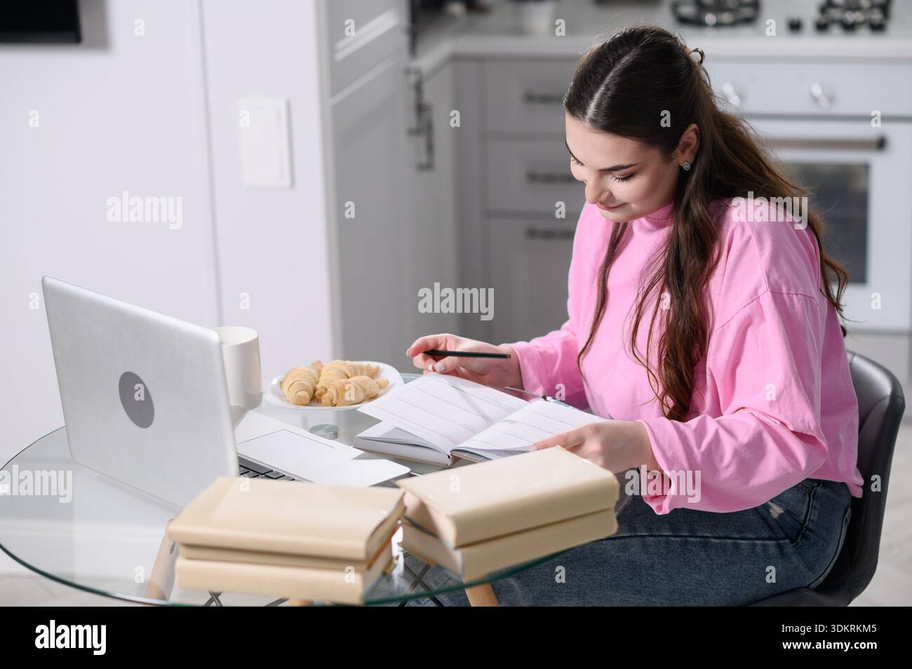 Woman organizes morning routine using laptop, daily planner, sitting at kitchen table with morning breakfast, coffee, croissants, creates daily schedule. Student prepares for exam surrounded by books Stock Photo