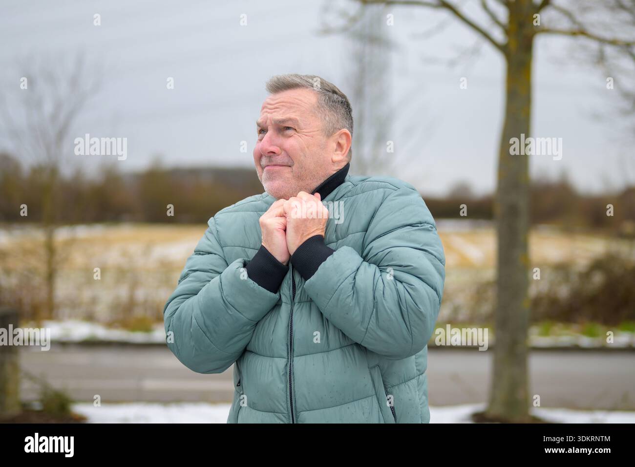 Mature man standing outdoors in winter, holding his jacket collar and looking into the distance with a thoughtful and slightly concerned expression ag Stock Photo