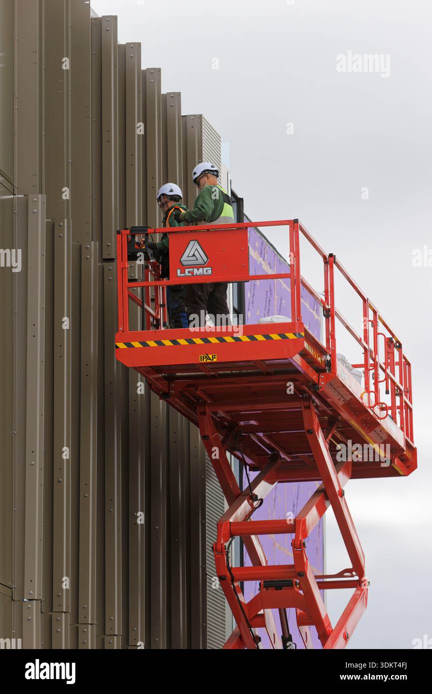 Two contractors with ppe on scissor lift fix aluminium fins to vertical ...