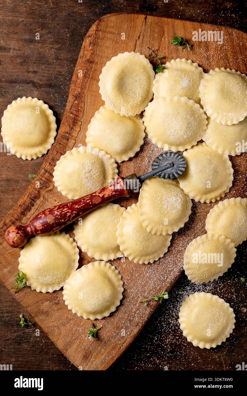 Fresh homemade ravioli pasta on wooden cutting board with vintage pasta cutter tool and herbs dusted with flour on rustic kitchen table Stock Photo