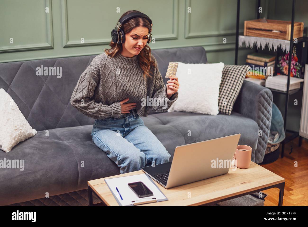 Young woman is sitting on bed in living room and having online doctor's appointment using laptop. Young woman taking medication while working on lapto Stock Photo