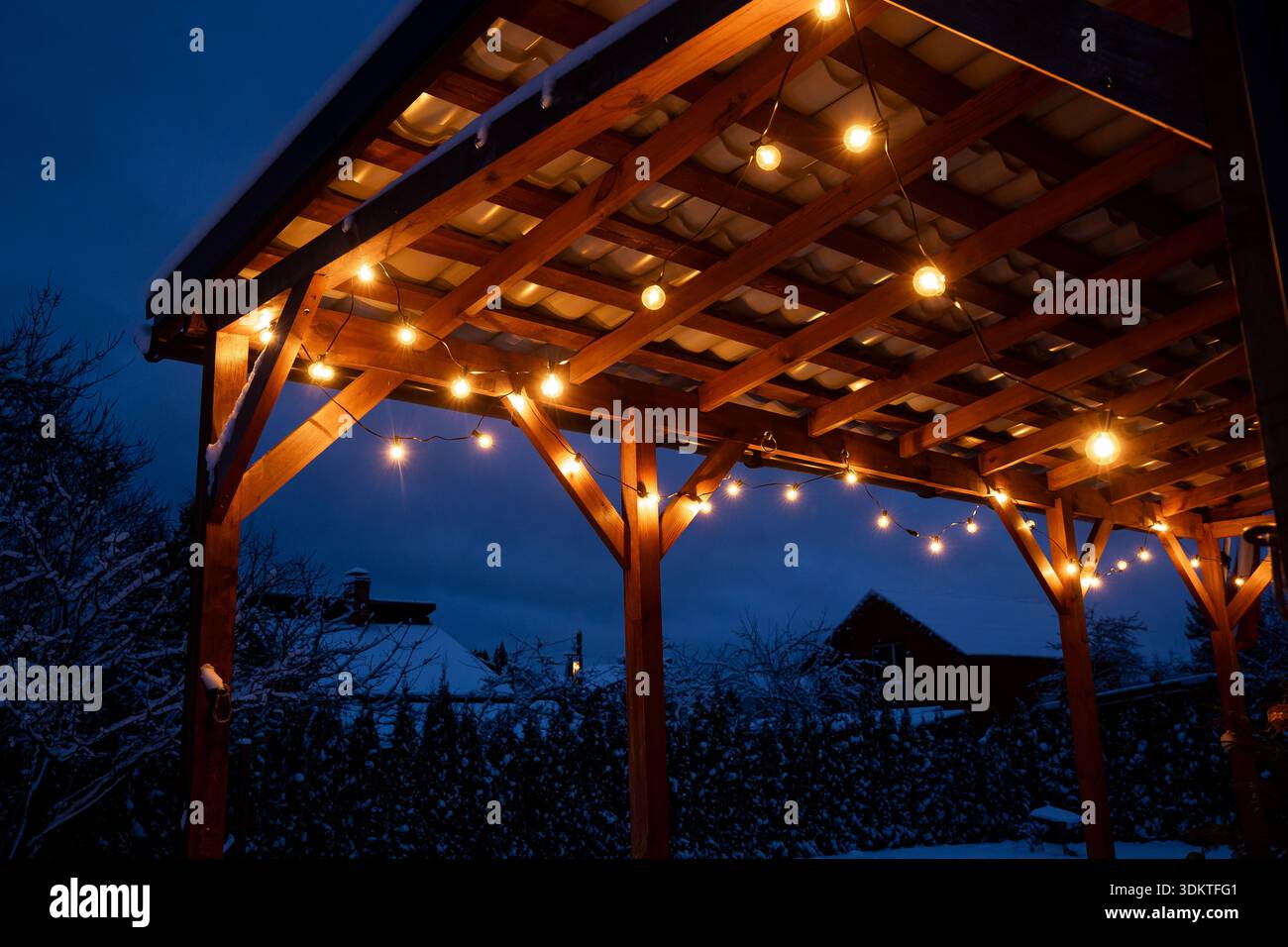 A wooden terrace illuminated by warm LED string lights, surrounded by snow covered trees and rooftops under a dark blue evening sky. Stock Photo