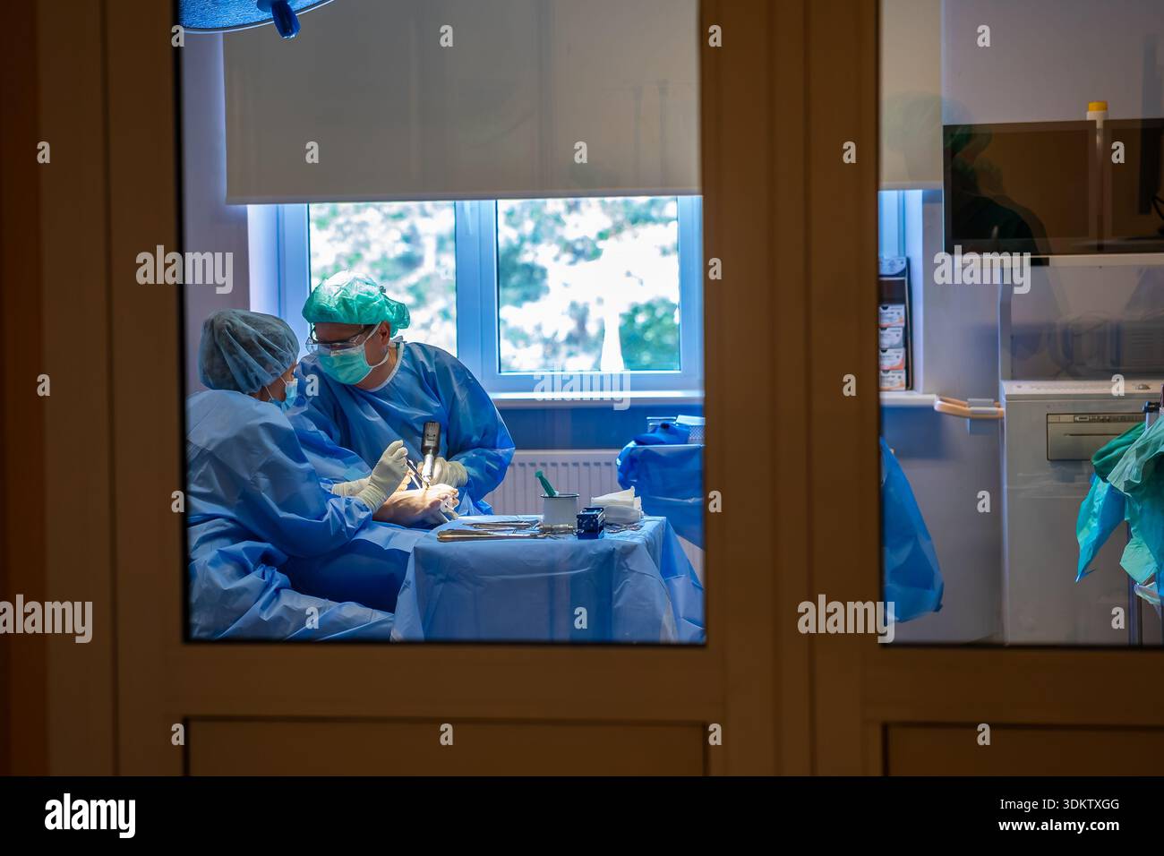 Two professionals in blue surgical attire work in a well lit operating room with visible equipment, cabinets, and a monitor, viewed through a glass do Stock Photo