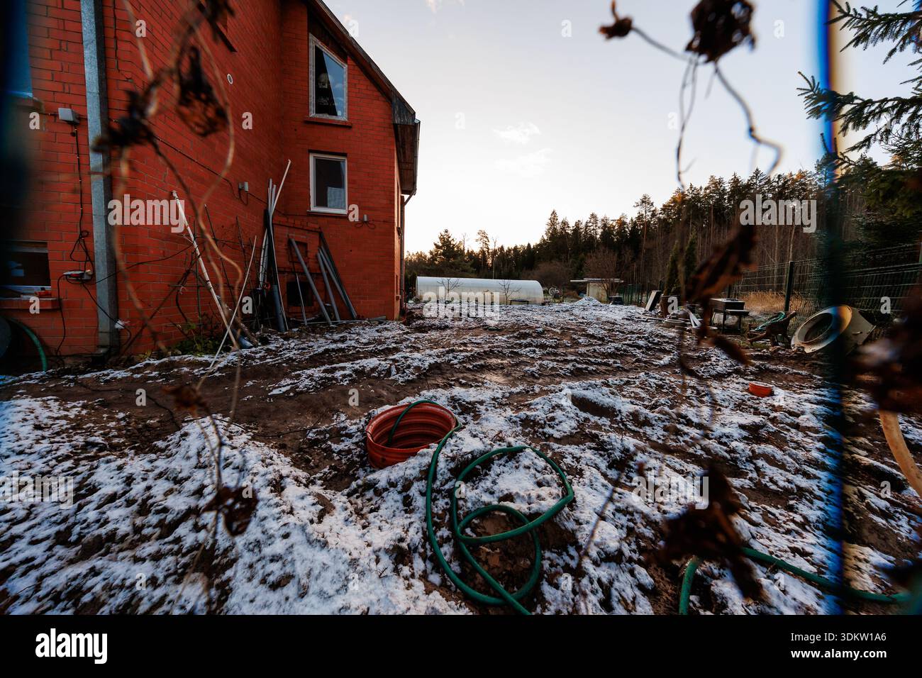 A red brick two story house borders a snow dusted yard with hoses, ladders, pipes, and a white cylindrical tank by trees, in late afternoon with long Stock Photo