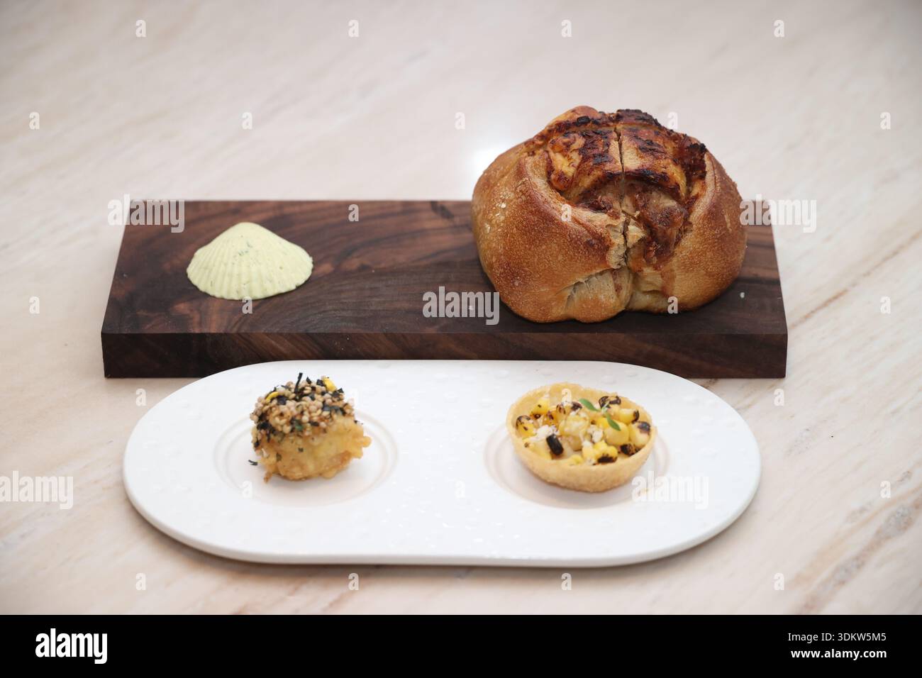Gourmet bread alongside delicate appetizers, including corn tartlet and fried scallop, served with smooth butter on a wooden board and white platter Stock Photo
