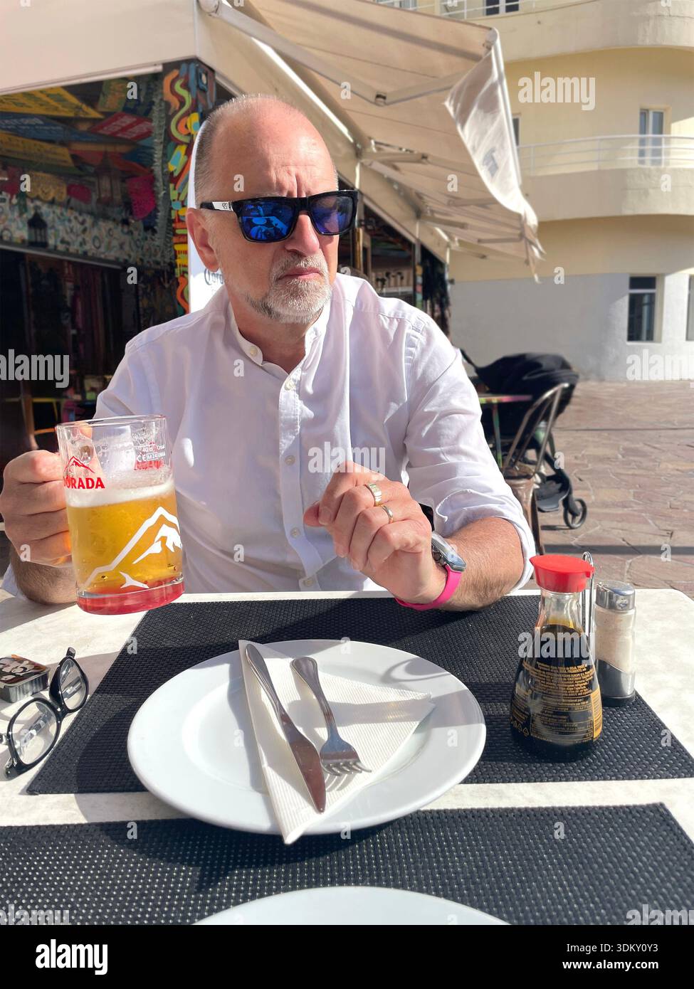 A middle-aged man sits at an outdoor café table with a beer, looking pensive while enjoying a meal spain - Smartphone Captured Stock Image