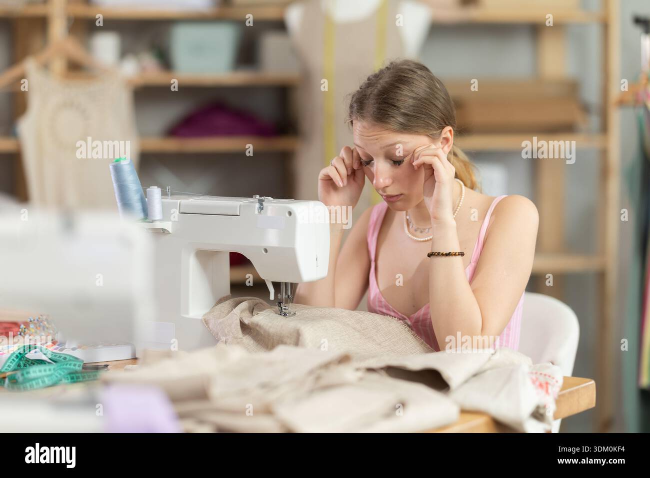 Tired young tailoress sitting at the sewing-machine in tailor's shop ...
