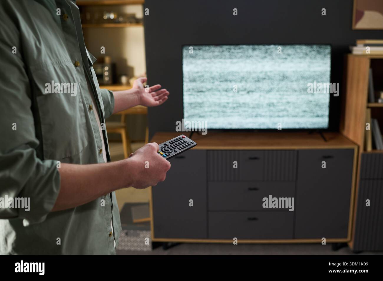 Midsection shot of man standing in living room holding remote control gesturing toward television displaying static screen, showing frustration with malfunctioning device Stock Photo
