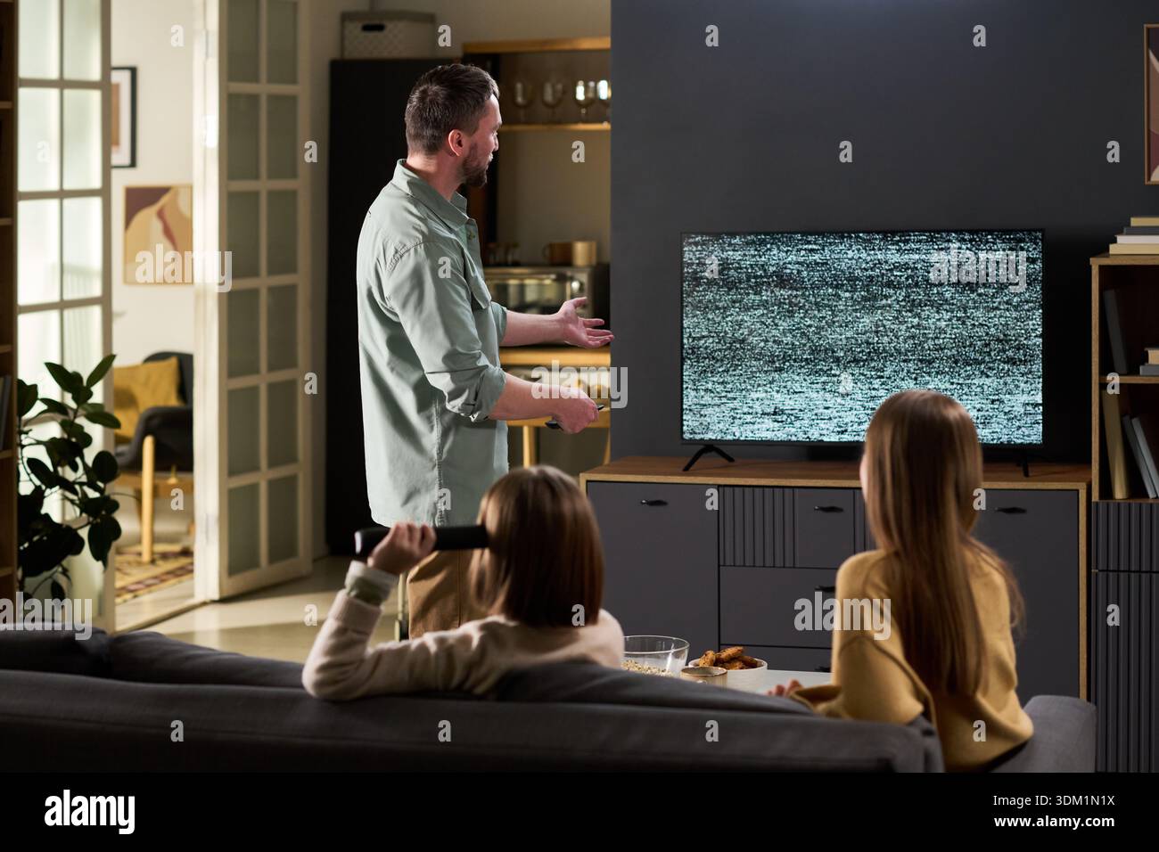 Caucasian man standing near television with static screen gesturing while two Caucasian wife and child sitting on sofa holding remote control watching television together Stock Photo