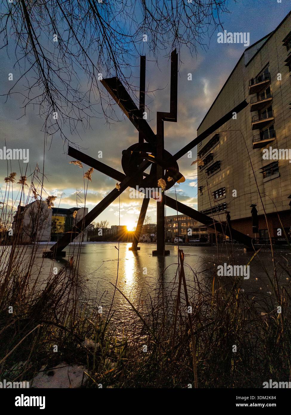 Dramatic sunset view of 'Galileo' by Mark di Suvero sculpture  casting intricate reflections on the water amid golden-hour glow and surrounding highri - Smartphone Captured Stock Image