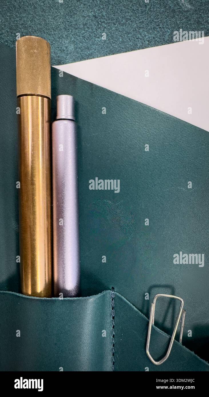 Brass and rose gold pens in leather pouch on emerald green notebook with paper clip, minimalist desk setup. - Smartphone Captured Stock Image