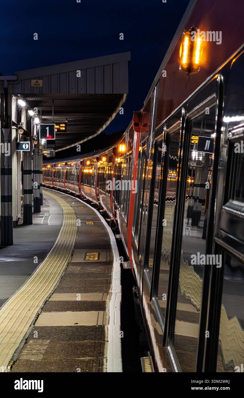 Red southern rail commuter train at night on curved platform with glowing yellow headlights and platform lights, modern canopy structure, tactile paving, and reflection - Smartphone Captured Stock Image