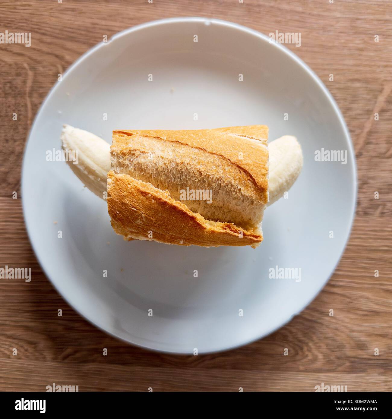 Novelty banana-stuffed baguette sandwich centered on white plate over wooden table, playful food concept with curved banana protruding from crusty Fre - Smartphone Captured Stock Image