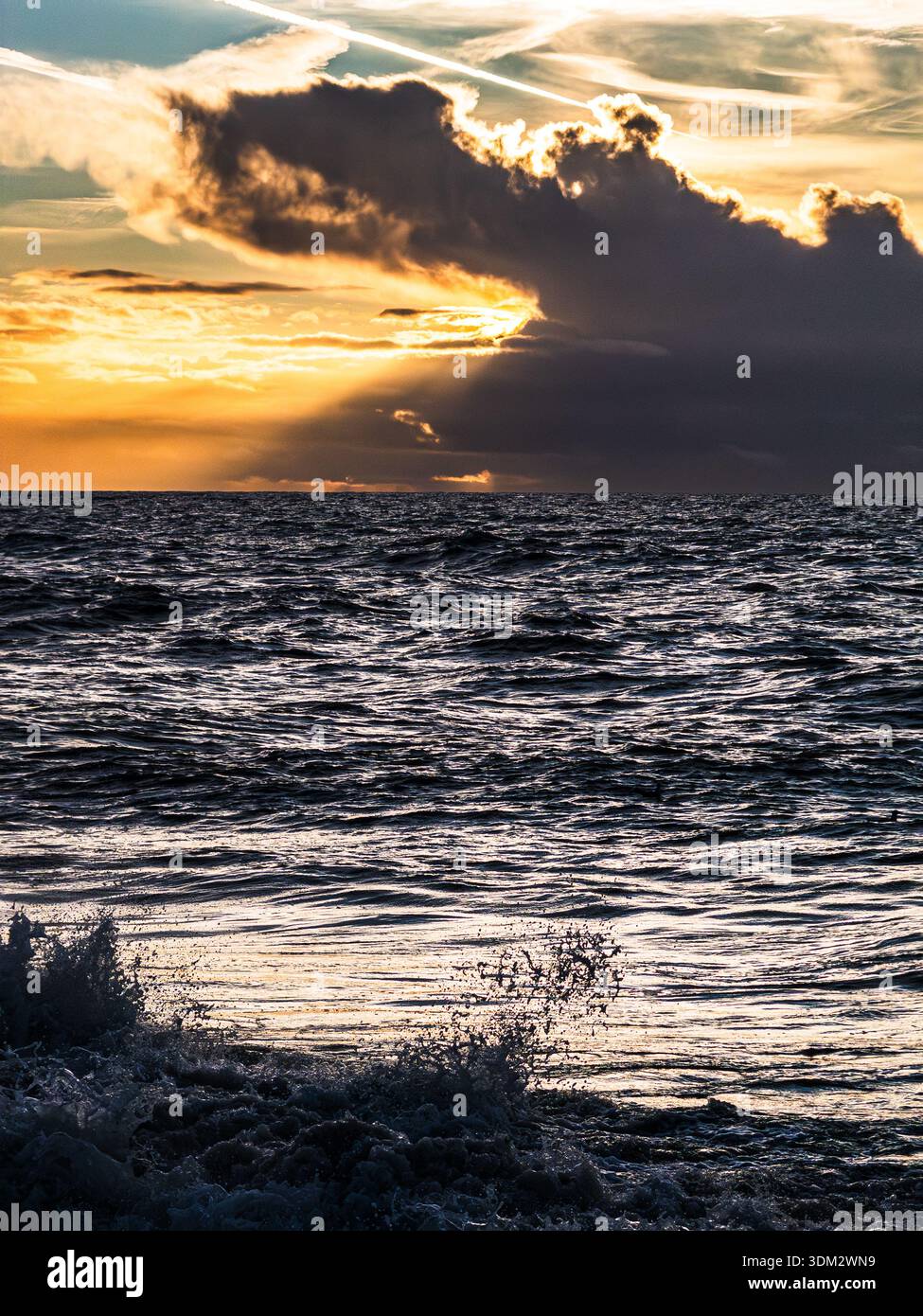 Epic sunrise over English Channel from Worthing beach with dramatic orange-glowing cloud pierced by airplane contrail and golden sun rays beaming towa - Smartphone Captured Stock Image