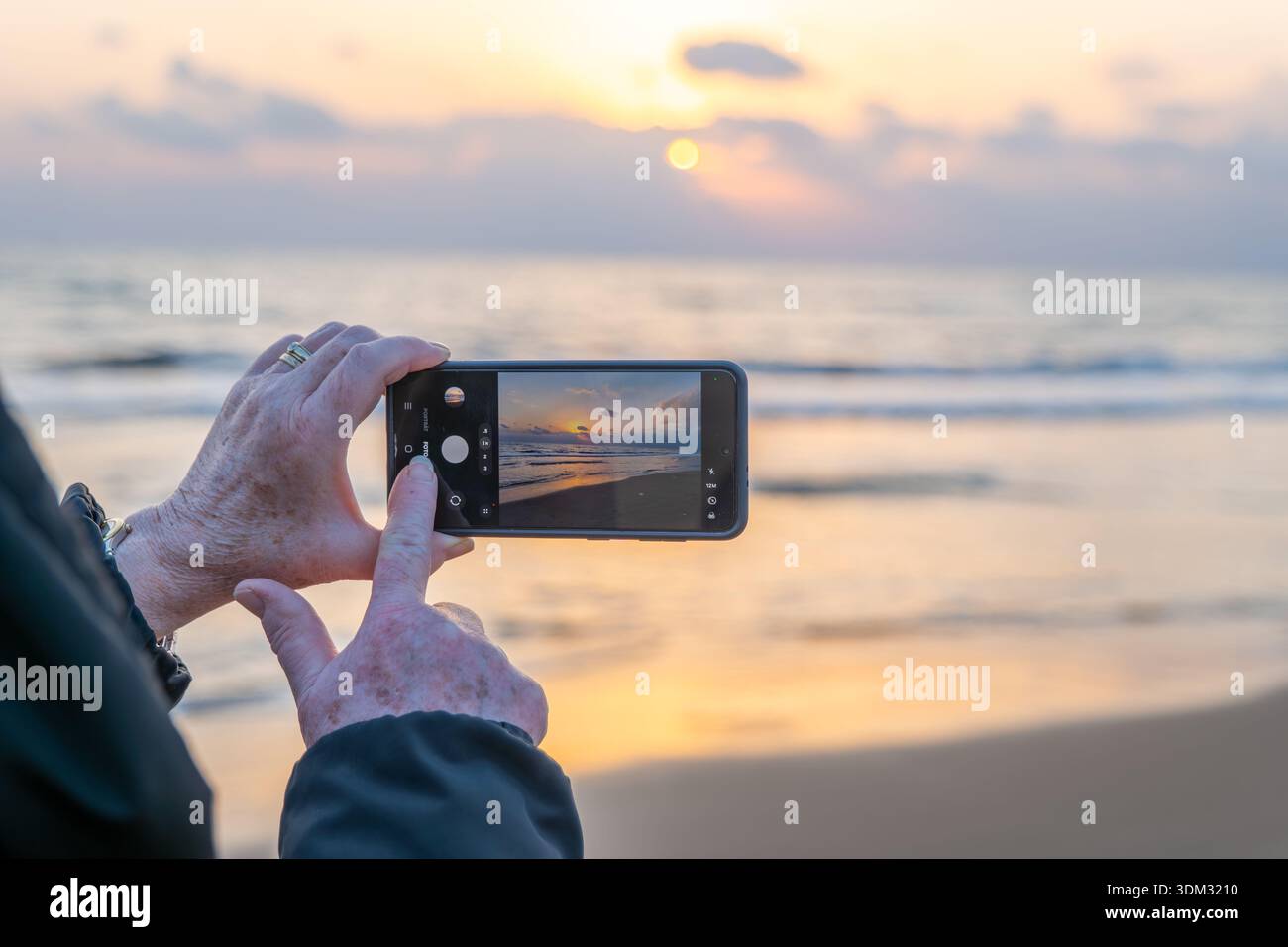 A person using a mobile phone to take a picture of a dramatic sunset at the beach, with a finger touching the shutter button on the screen. Stock Photo