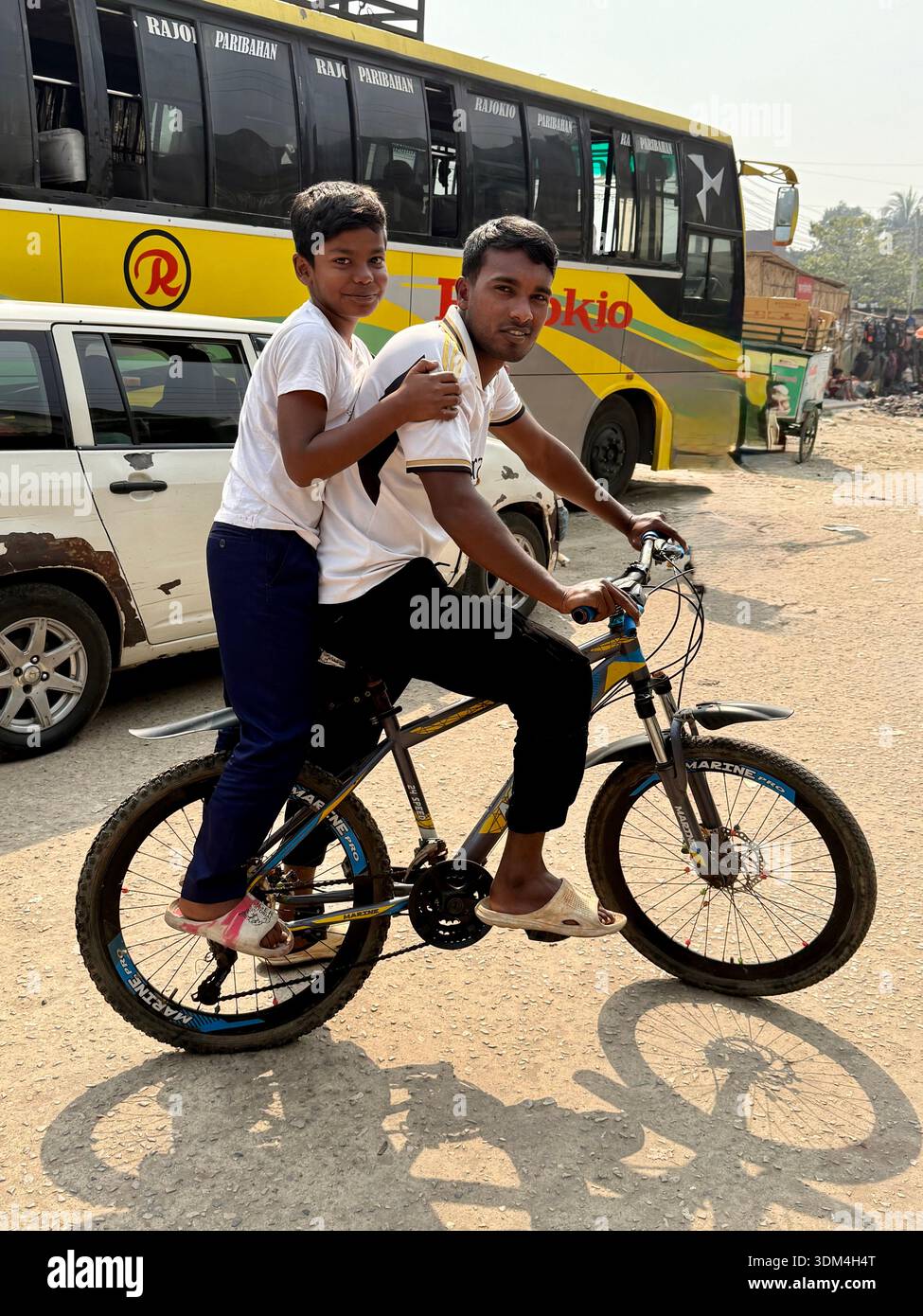 oung boy riding as a passenger on a bicycle with an adult man along a dusty roadside in Dhaka, Bangladesh. - Smartphone Captured Stock Image