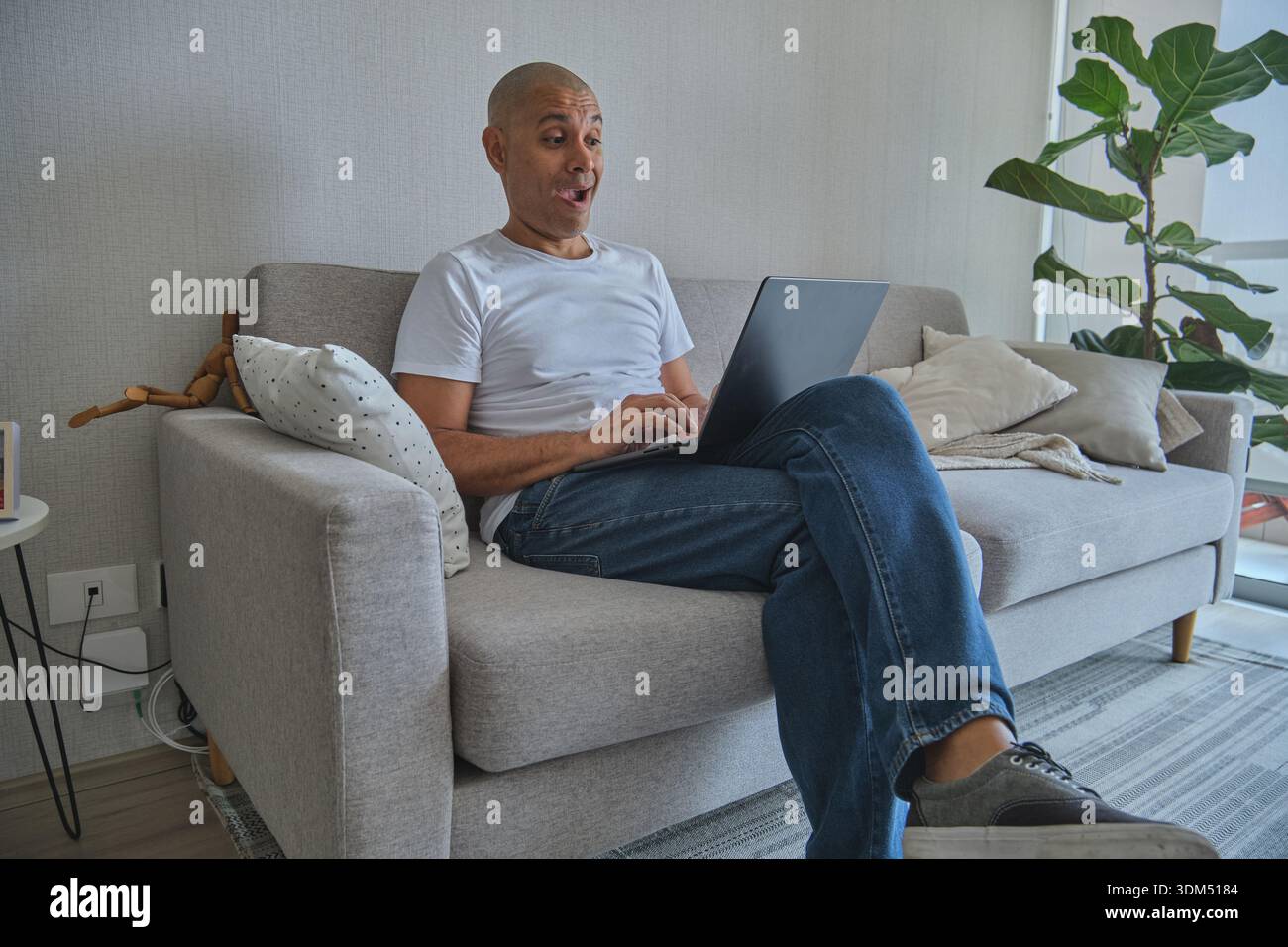 Middle-aged man sitting on sofa, reacting with surprise to content on his laptop screen Stock Photo