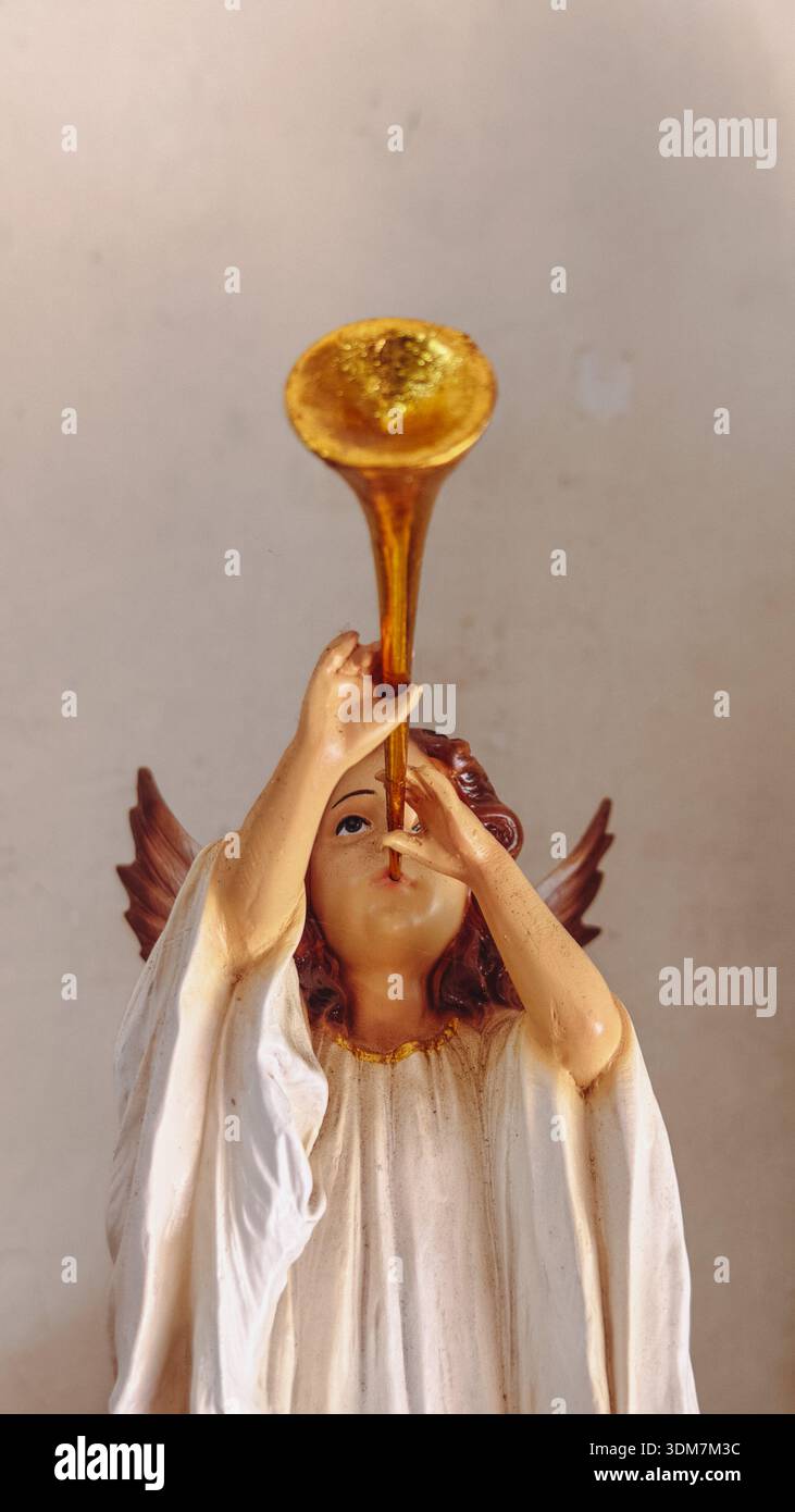 Angel figurine blowing golden trumpet, white robe, gold necklace, brown hair, wings, plain background. Stock Photo