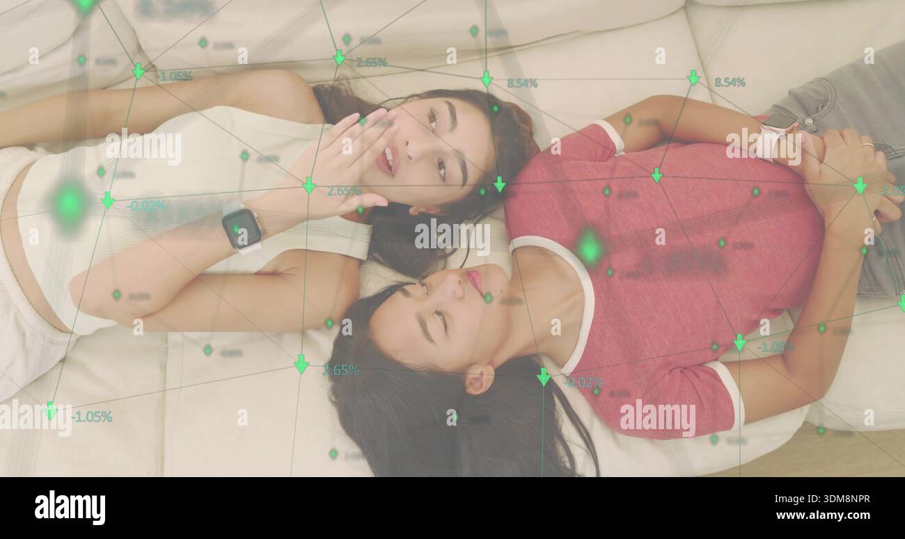 Lying two women talking on sofa cushions, wearing smartwatches, crop top, red tee, green overlay Stock Photo