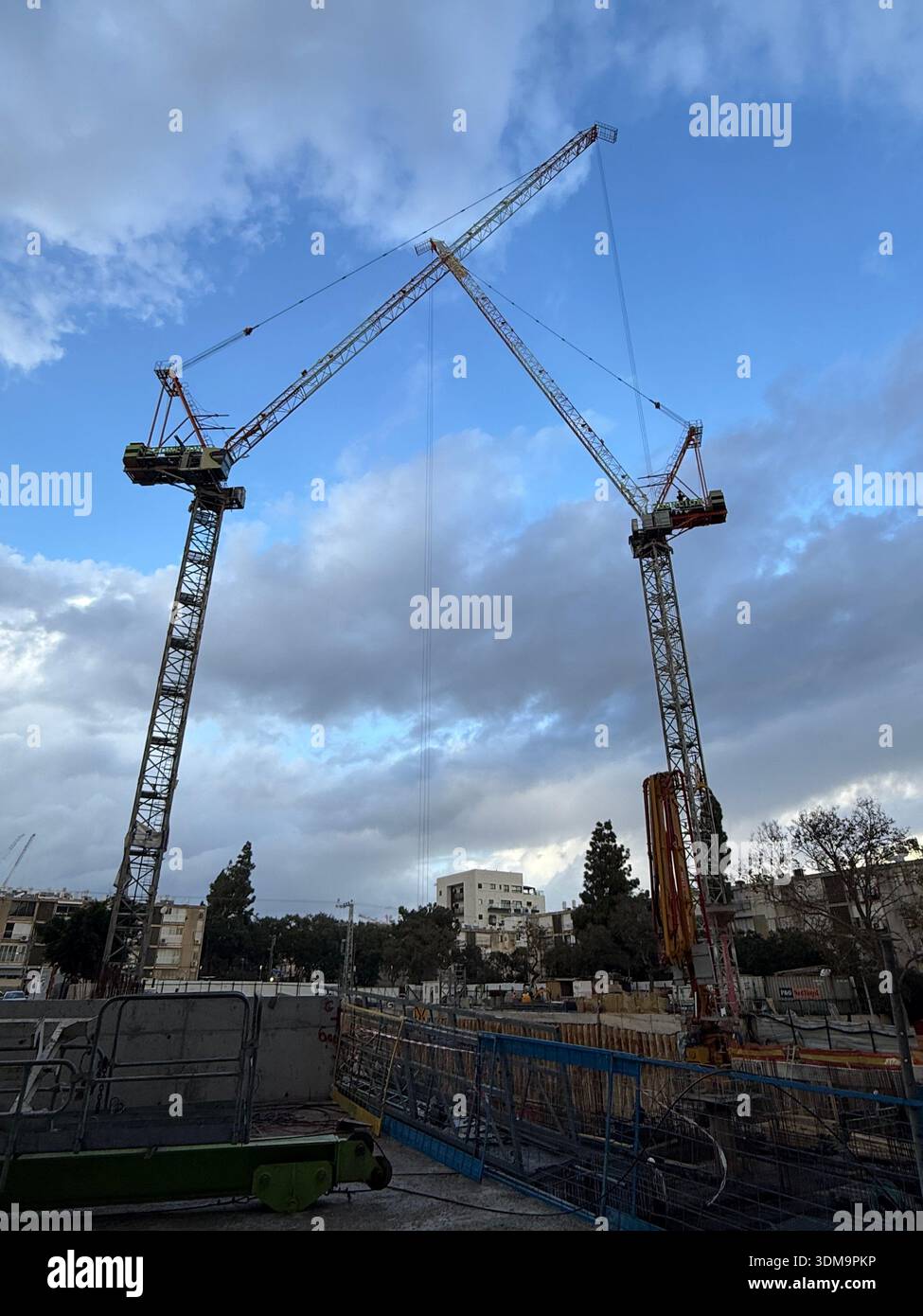 Two towering construction cranes reach toward a cloudy blue sky over a busy urban building site. Industrial equipment and city structures frame the sc - Smartphone Captured Stock Image