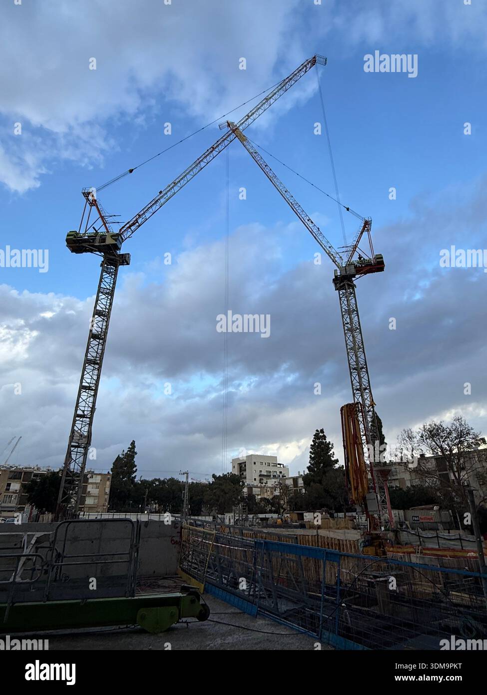 Two towering construction cranes reach toward a cloudy blue sky over a busy urban building site. Industrial equipment and city structures frame the sc - Smartphone Captured Stock Image