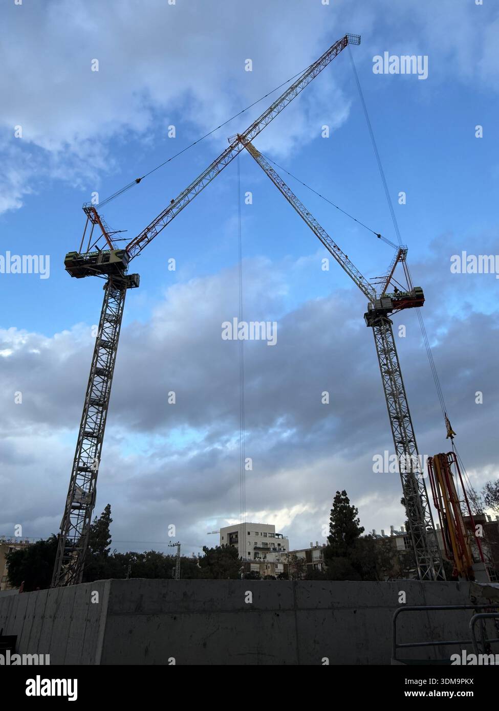 Two towering construction cranes reach toward a cloudy blue sky over a busy urban building site. Industrial equipment and city structures frame the sc - Smartphone Captured Stock Image