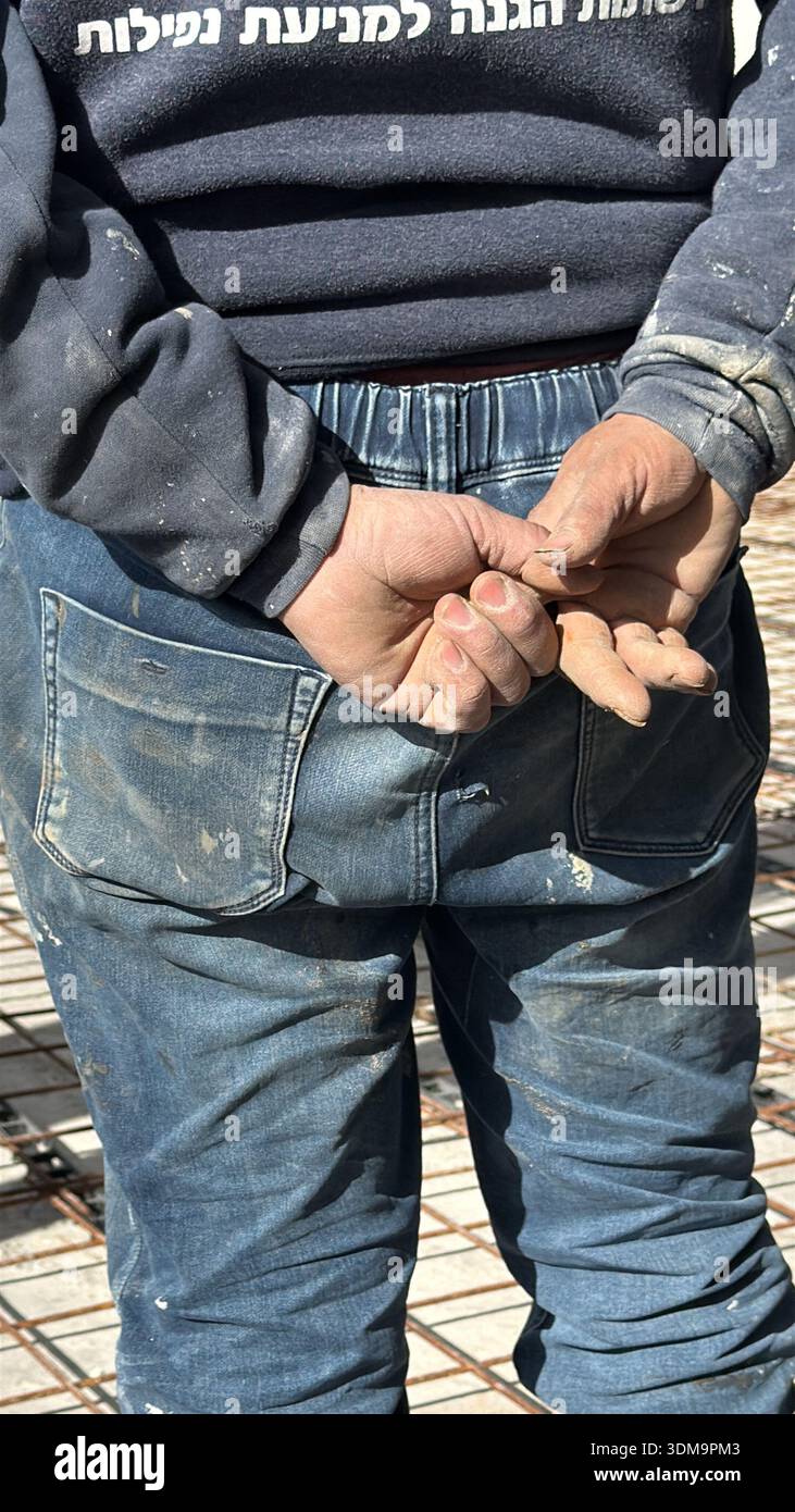 A worker's dusty hands are clasped behind their back, wearing paint-splattered jeans on a rebar construction site. A grit-and-toil urban close-up. - Smartphone Captured Stock Image