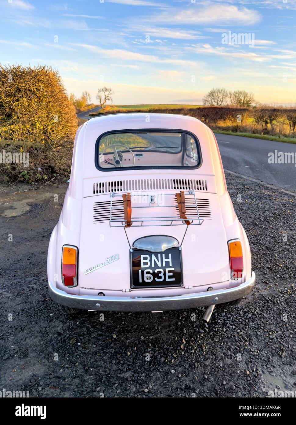 A vintage pink classic Fiat 500 car parked on a rural road, surrounded by a serene landscape at sunset - Smartphone Captured Stock Image