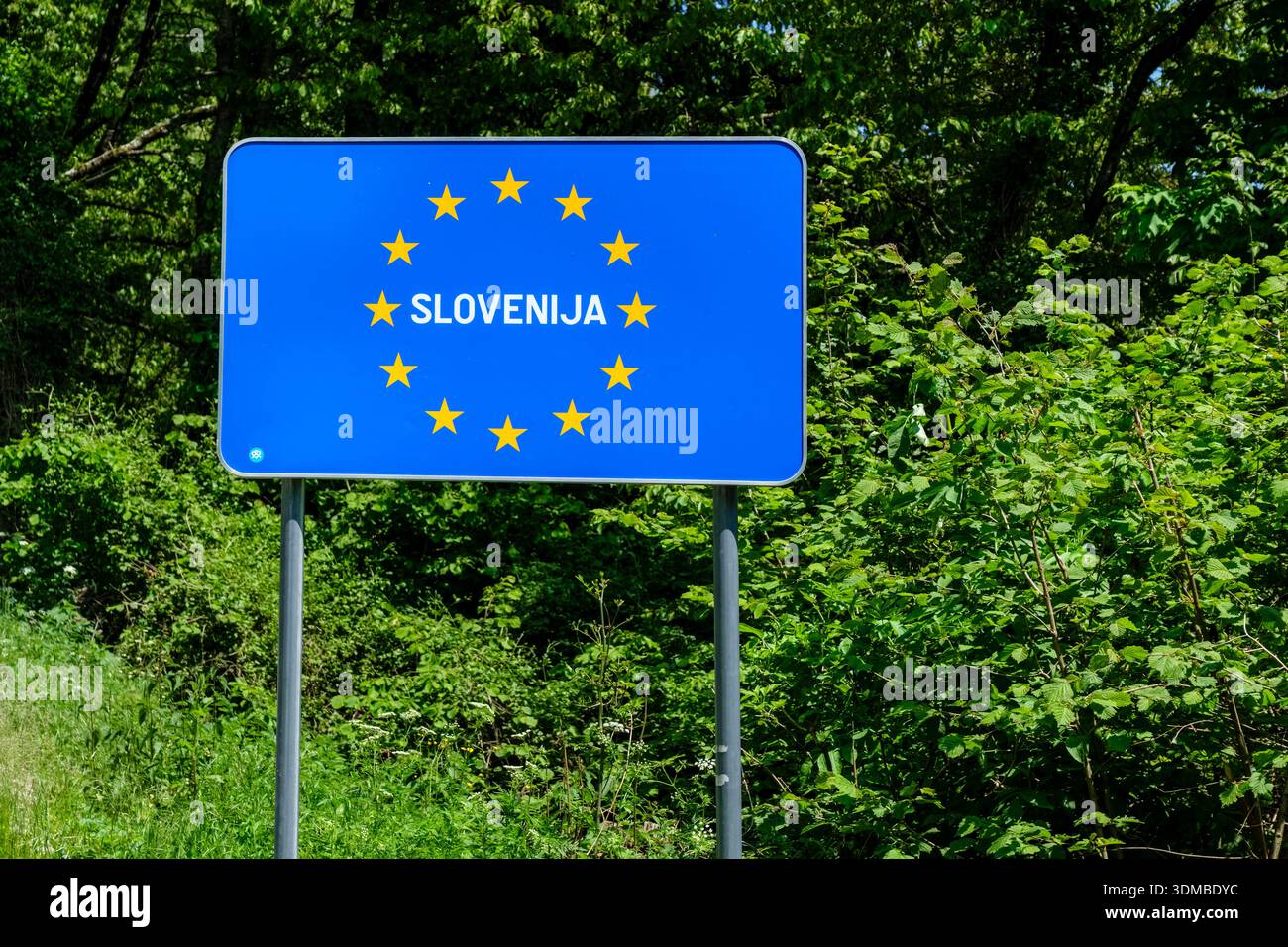 Blue European Union border sign for Slovenia featuring yellow stars, standing in front of green foliage and trees. Stock Photo
