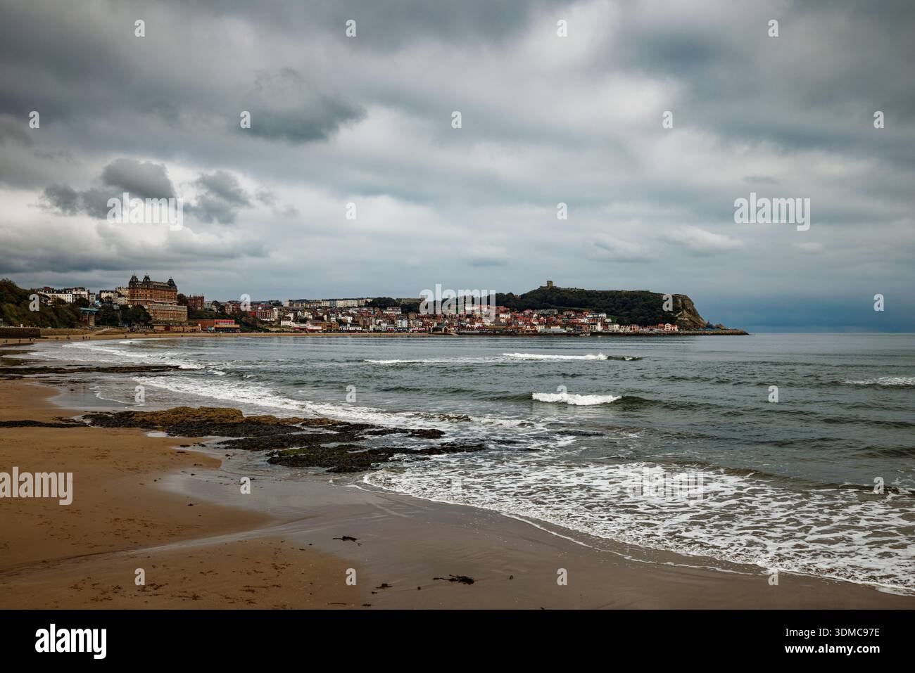 Gentle waves wash onto a beach on a cloudy day with the resort of Scarborough in the distance and castle on the headland, North Yorkshire. Stock Photo