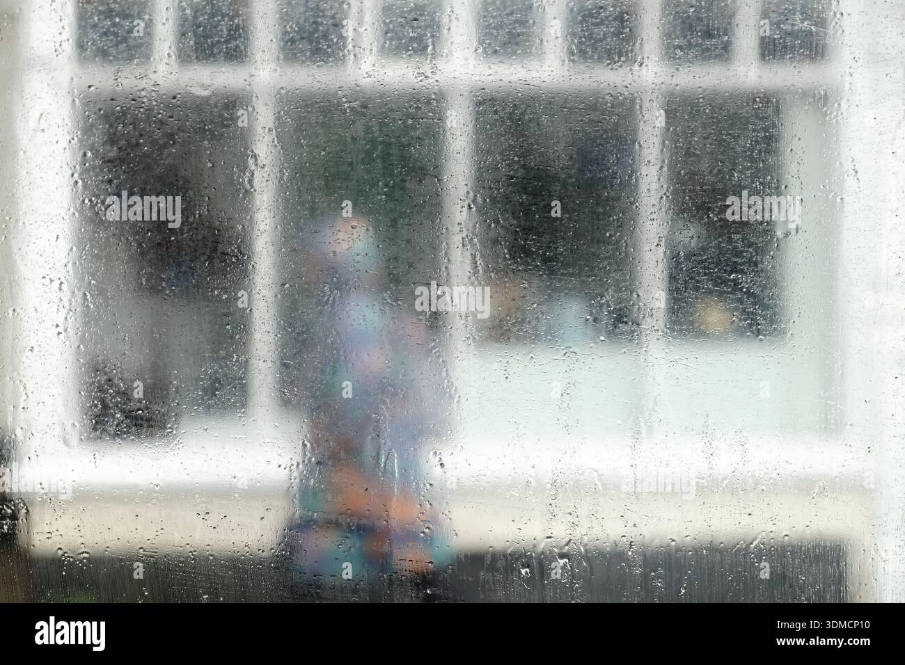 A person in a rain coat walking past a shop front in wet weather seen through a rain soaked window on a damp wet gloomy winter day. UK Stock Photo