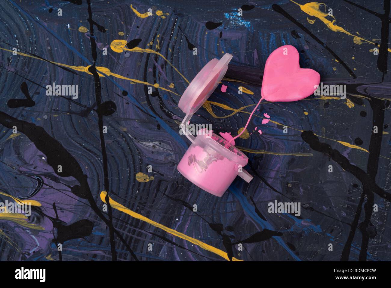 Vibrant flat lay featuring miniature pink heart emerging from bright pink paint pot amidst swirling galaxy-inspired background with gold accents. Stock Photo