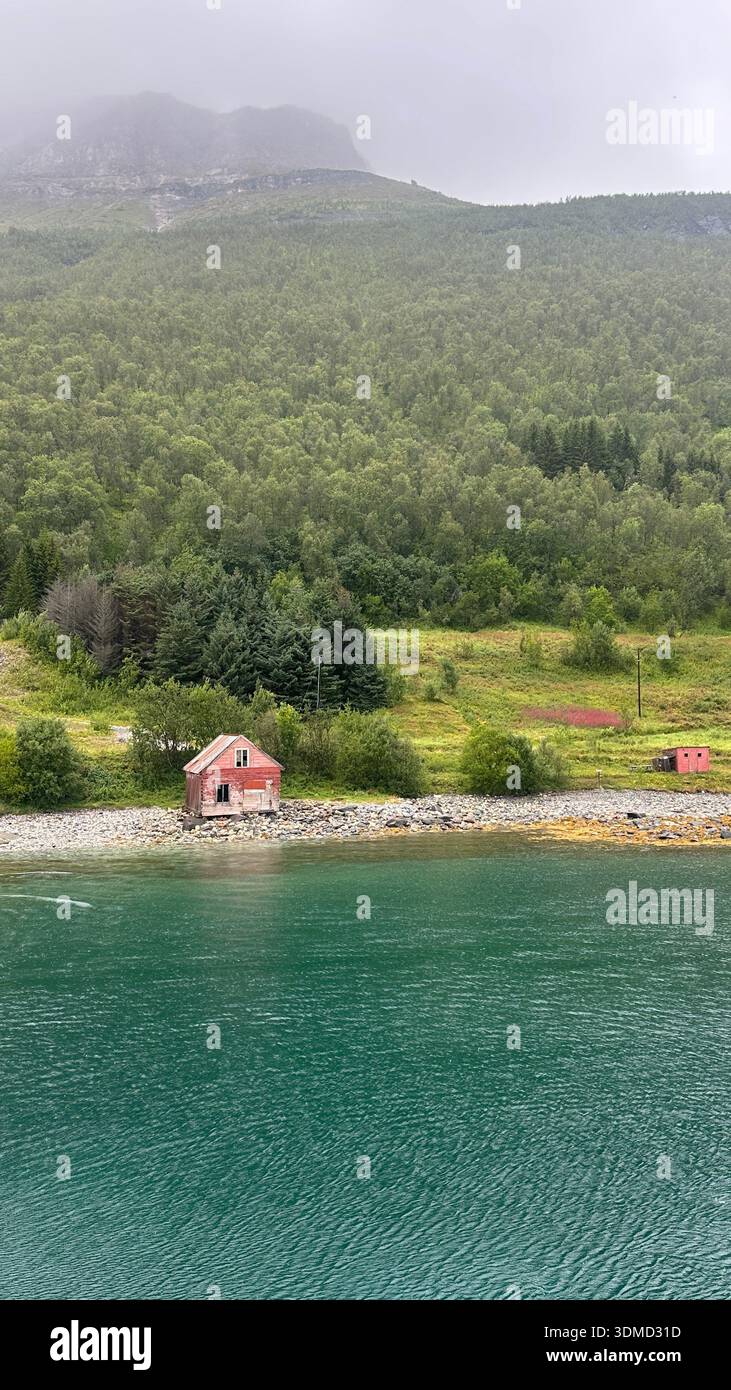 Small isolated red wooden house by the shoreline of a Norwegian fjord near Tromsø in Northern Norway. Arctic landscape with green hills and water - Smartphone Captured Stock Image