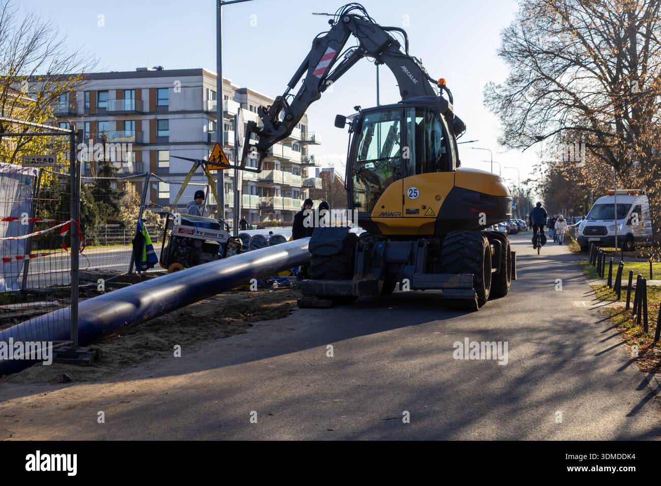 Poznan, Poland - November 21, 2025: Roadside scene of a yellow construction excavator and workers installing a large blue pipe for essential urban uti Stock Photo