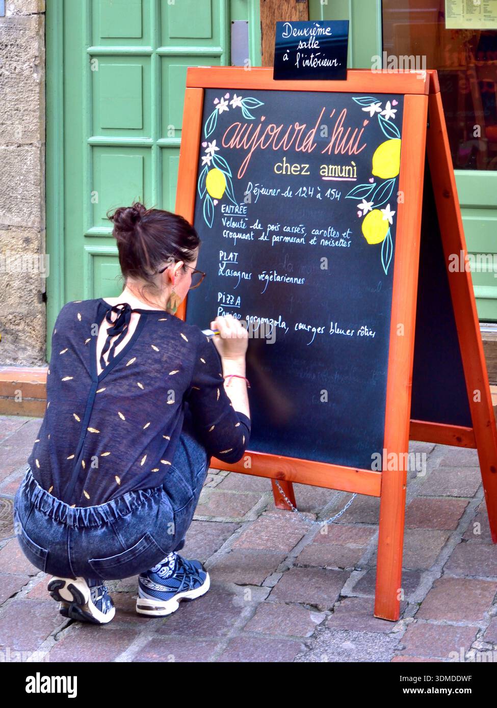 Young woman kneeling whilst writing the daily menu on a blackboard outside a restaurant - Tours, Indre-et-Loire (37), France. Stock Photo