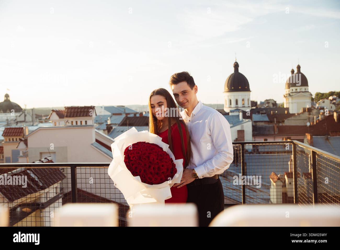 A couple stands on a rooftop holding a big bouquet of red roses while enjoying the view of the city at sunset. Stock Photo